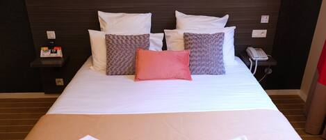 Una cama bien hecha con almohadas blancas y estampadas, una mesita de noche con un teléfono y una lámpara, y una lámpara fijada a la pared.