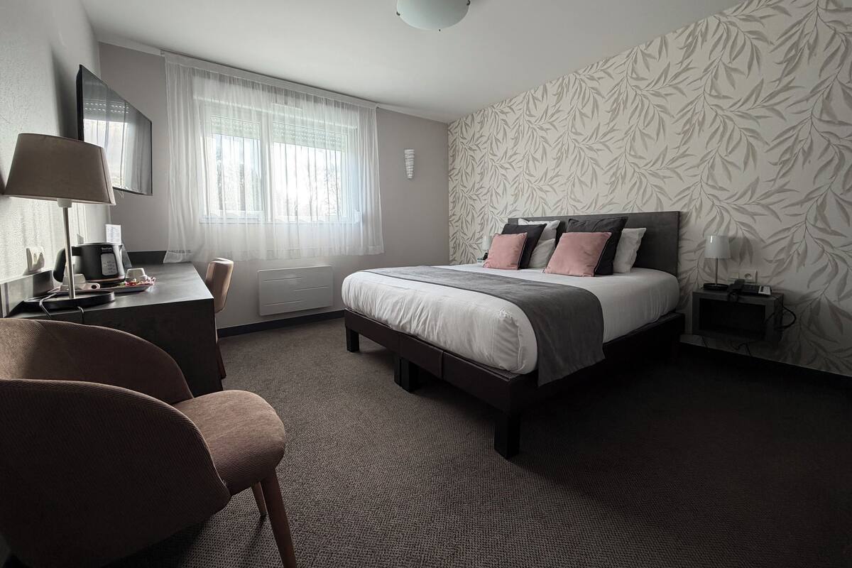 A modern hotel room with a bed, a desk, a chair, a TV, and a window with sheer curtains.