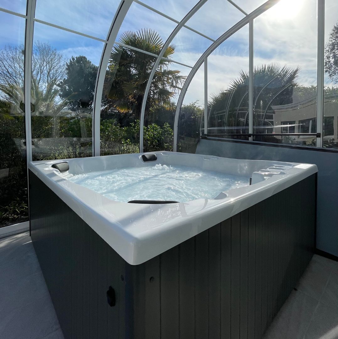 A hot tub with a glass enclosure, surrounded by greenery and a clear sky.