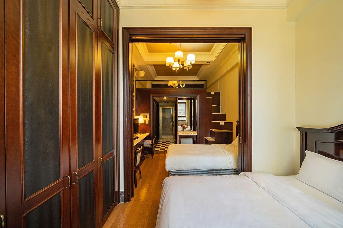 Quarto de hotel com duas camas, uma escrivaninha e uma cadeira. Dá para um corredor que leva a um banheiro e a uma escada.