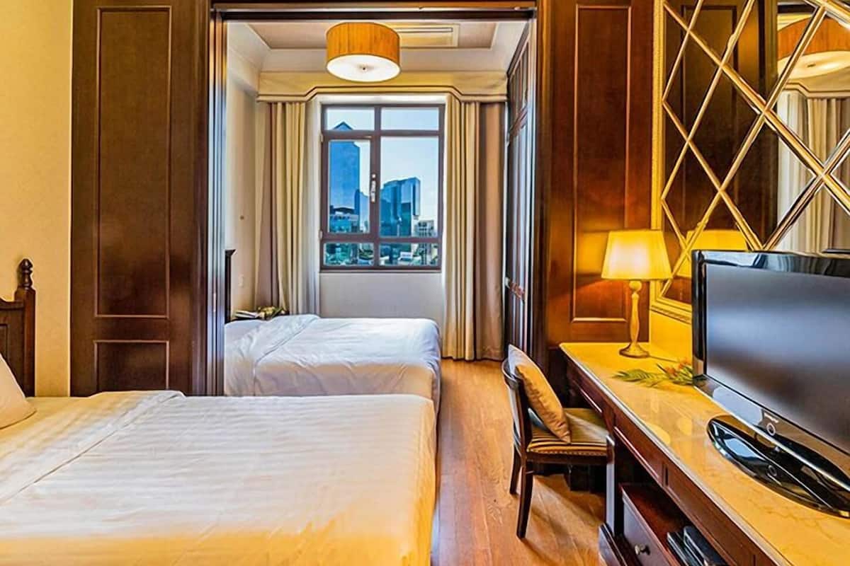 Quarto de hotel com duas camas, uma mesa de trabalho com uma televisão, janela com vista para a cidade e um candelabro.