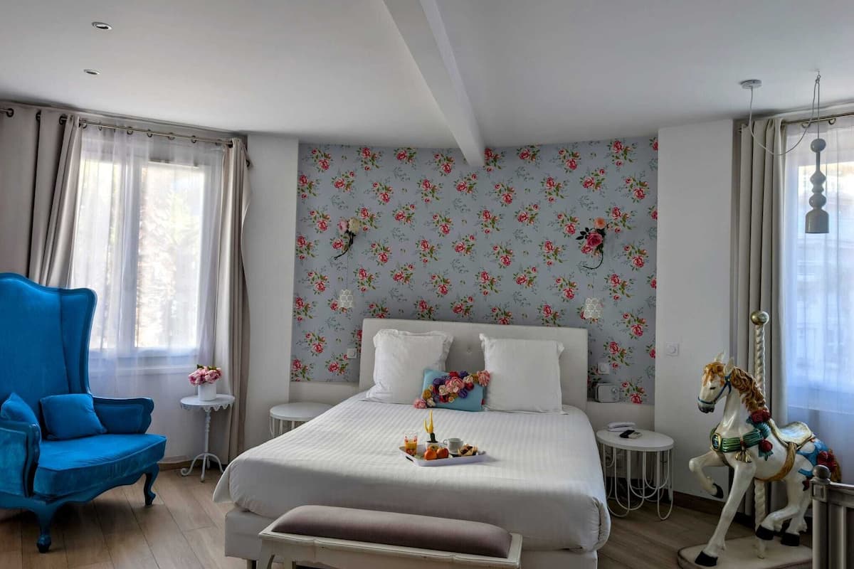A bedroom with a bed, a blue chair, a small round table, a decorative horse, and floral wallpaper.