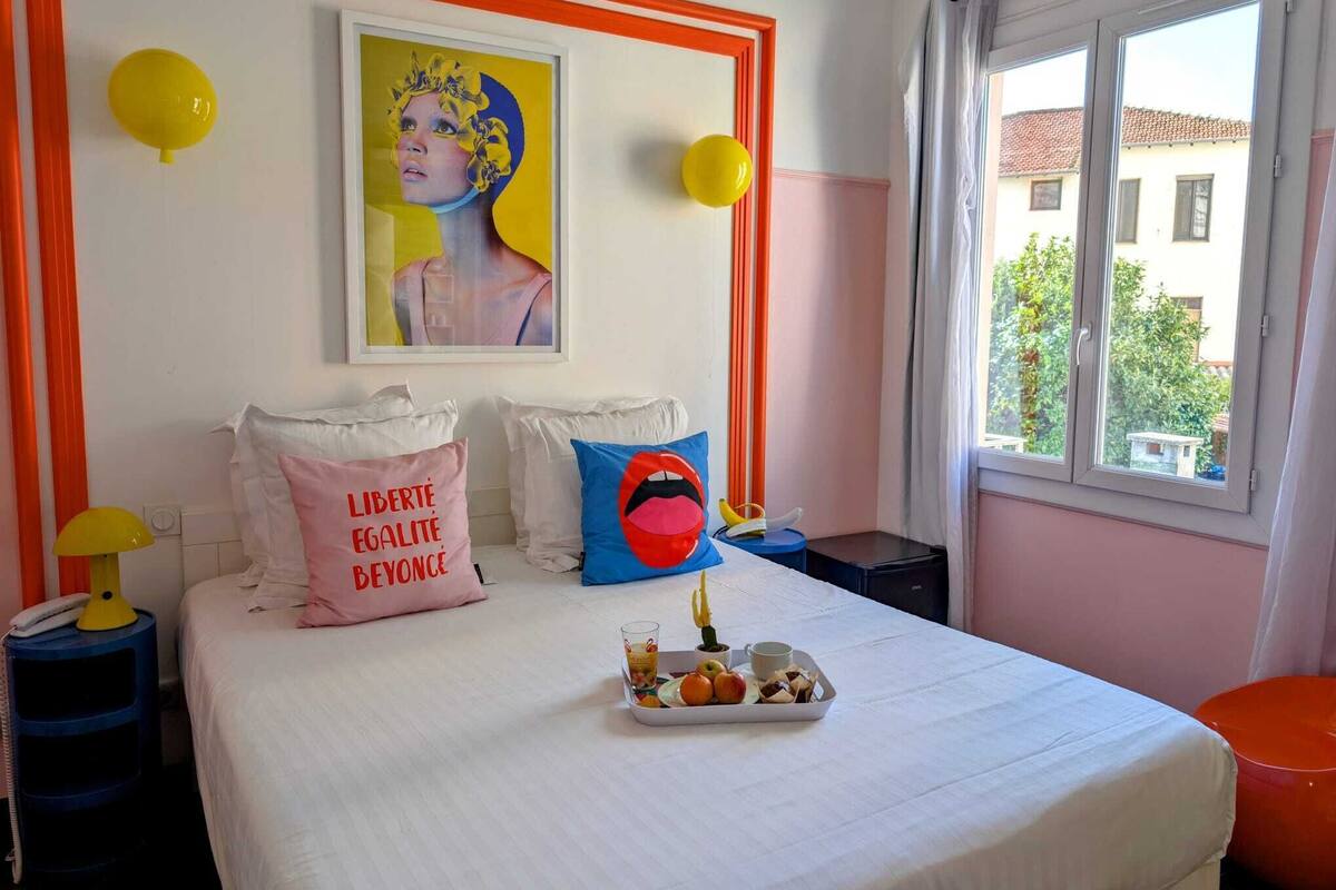 A bedroom with a bed, pillows, a tray of food, a painting, and a window with a view of buildings.