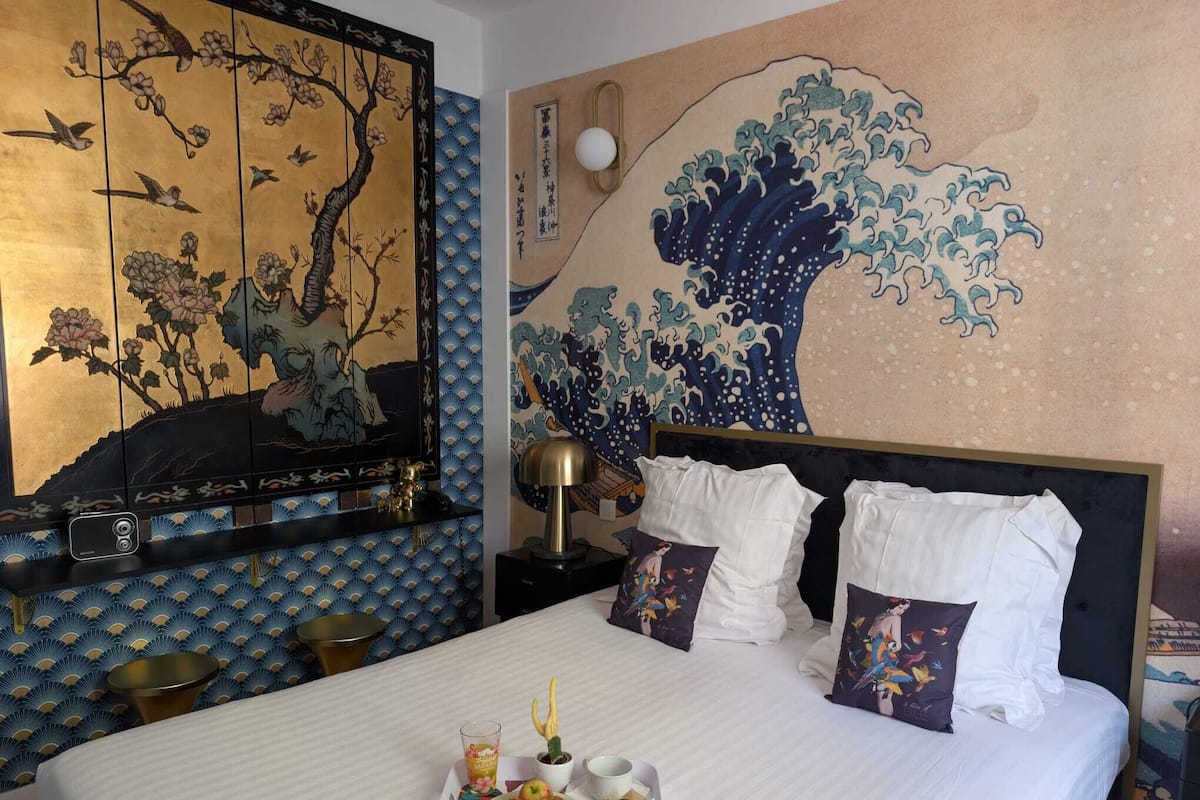 A hotel room with a traditional Japanese-style mural, a neatly made bed with white linens and decorative pillows, and a tray with fruits and a drink on the bed.