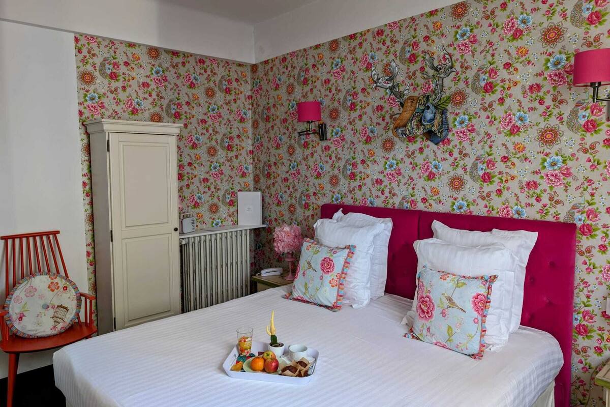 A bedroom with floral wallpaper, a pink headboard, a bedside table with a lamp, and a tray with fruits and a drink.