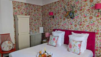 Un dormitorio con papel tapiz floral, cabecera rosa, mesita de noche con lámpara y una bandeja con frutas y una bebida.