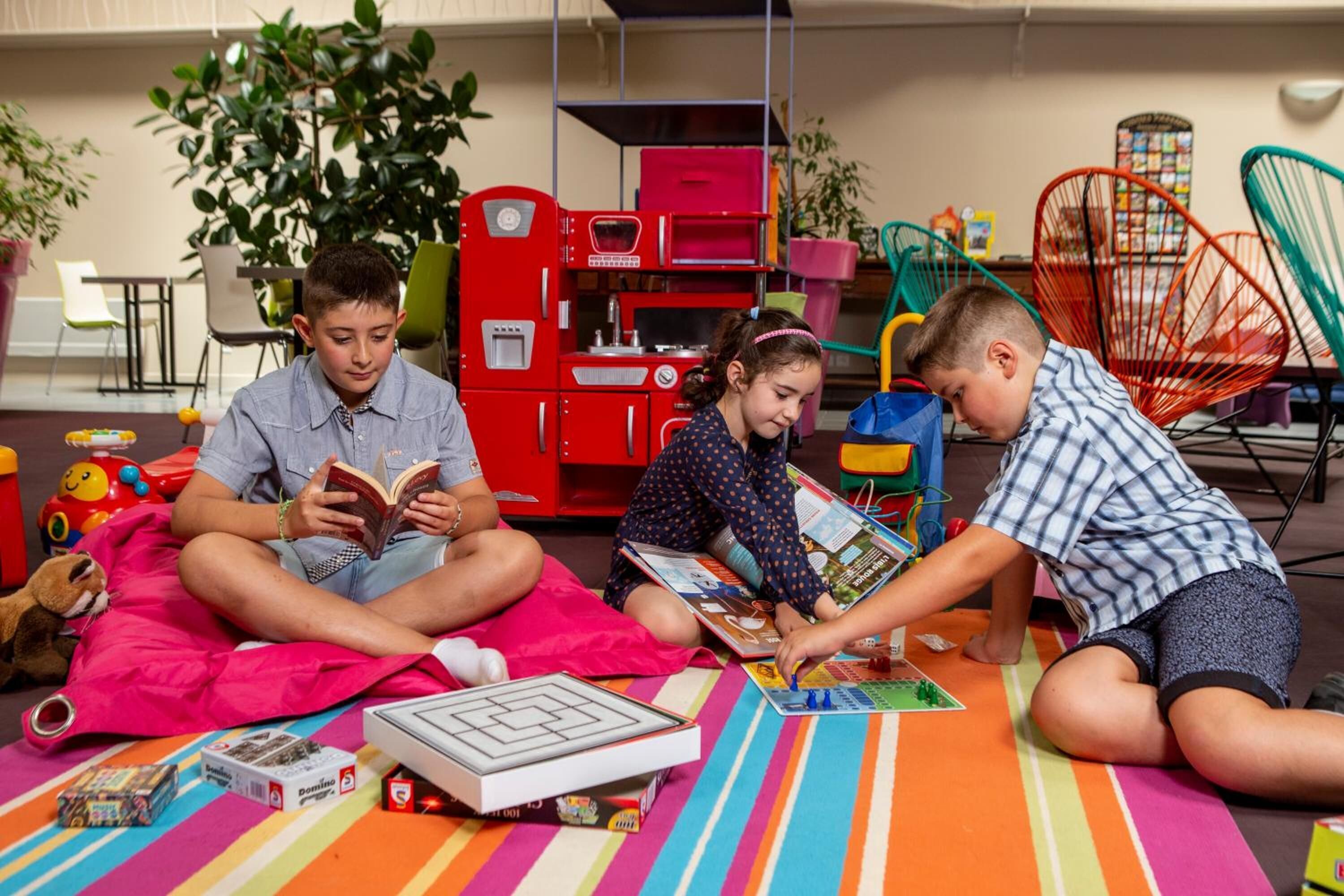 A group of children reading books in a brightly colored room with a red toy kitchen set and a large plant.