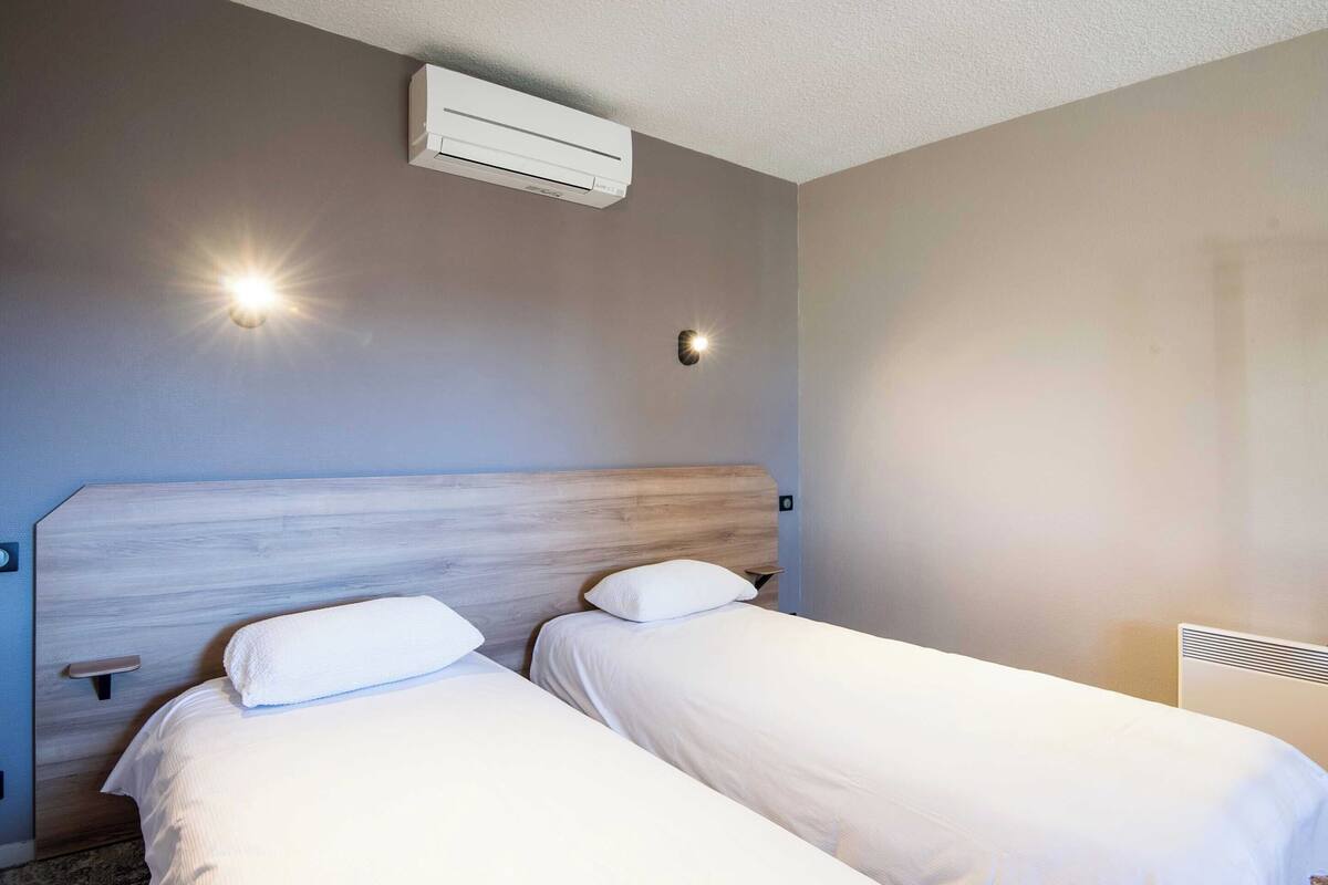 A hotel room with two beds, a wooden headboard, an air conditioning unit, and a wall-mounted light fixture.