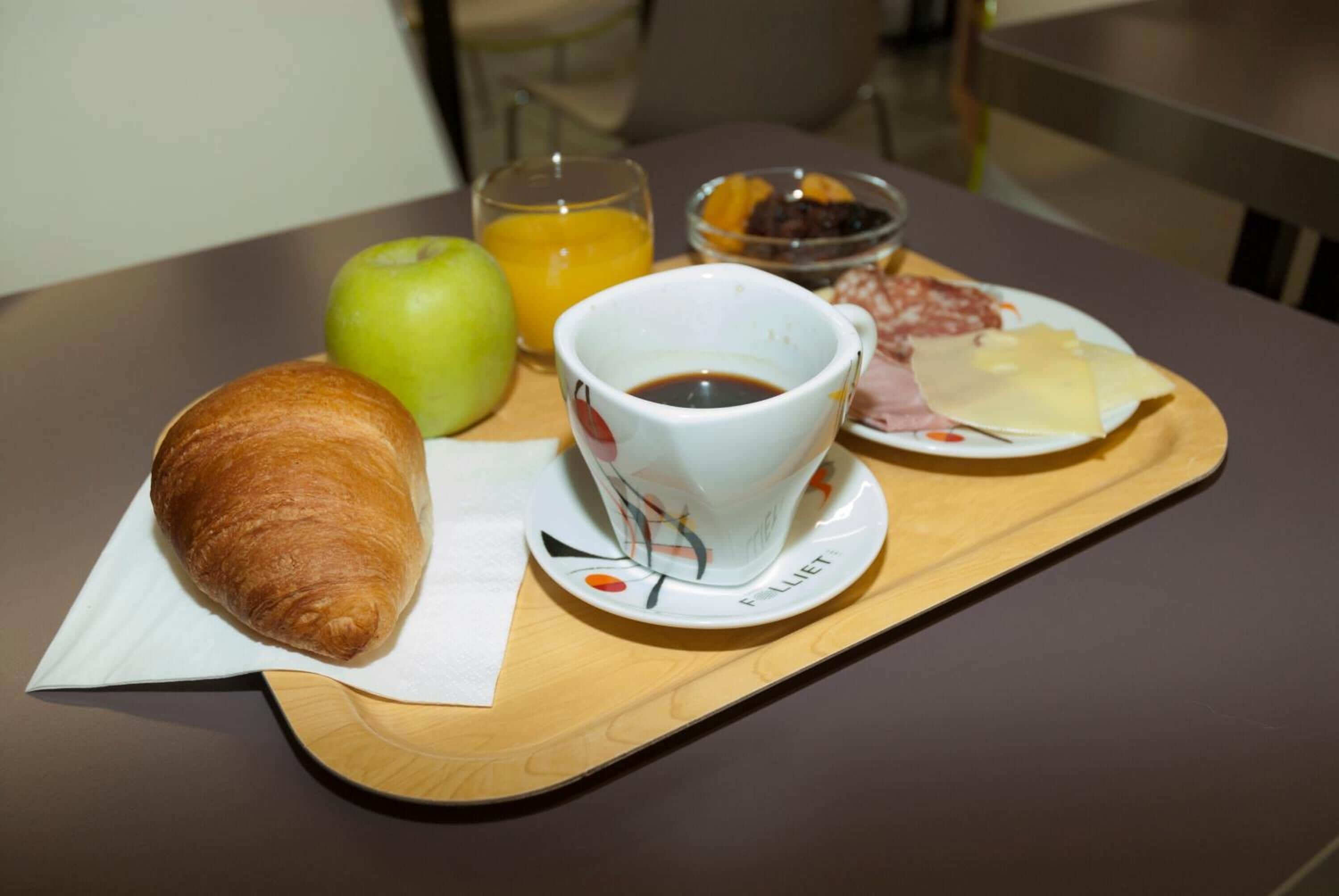 Bandeja de café da manhã com croissant, maçã, suco de laranja, xícara de café e um prato com queijo e salame.