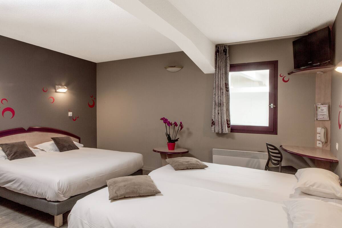 A hotel room with two beds, a small table with a red vase, a chair, a desk, a television, and a window with curtains.