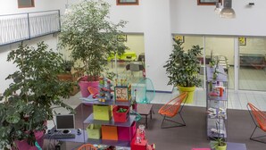 A modern indoor space with multiple seating areas, including colorful chairs and tables, surrounded by potted plants and shelves with various items.