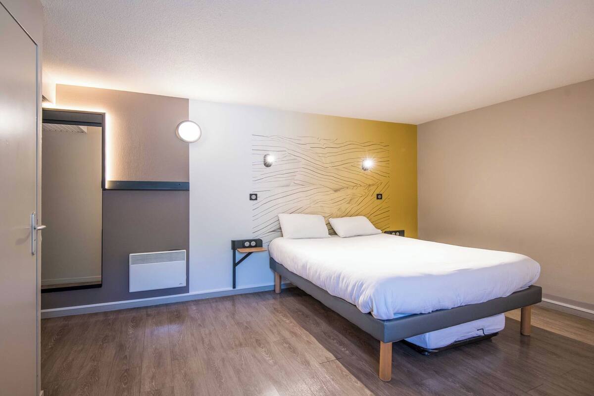 A modern bedroom with a bed, a nightstand, a wall-mounted heater, and a mirror.