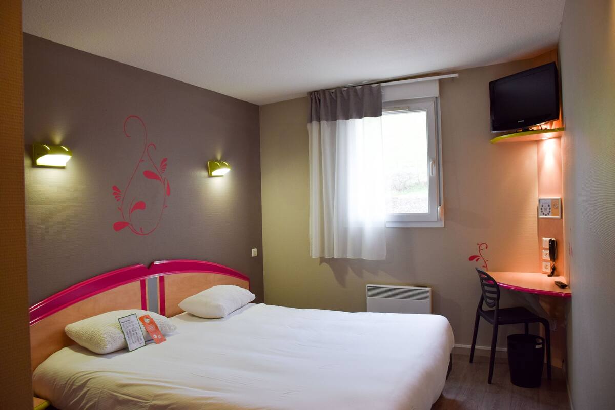 A hotel room with a bed, a desk, a chair, a TV, and a window with curtains.