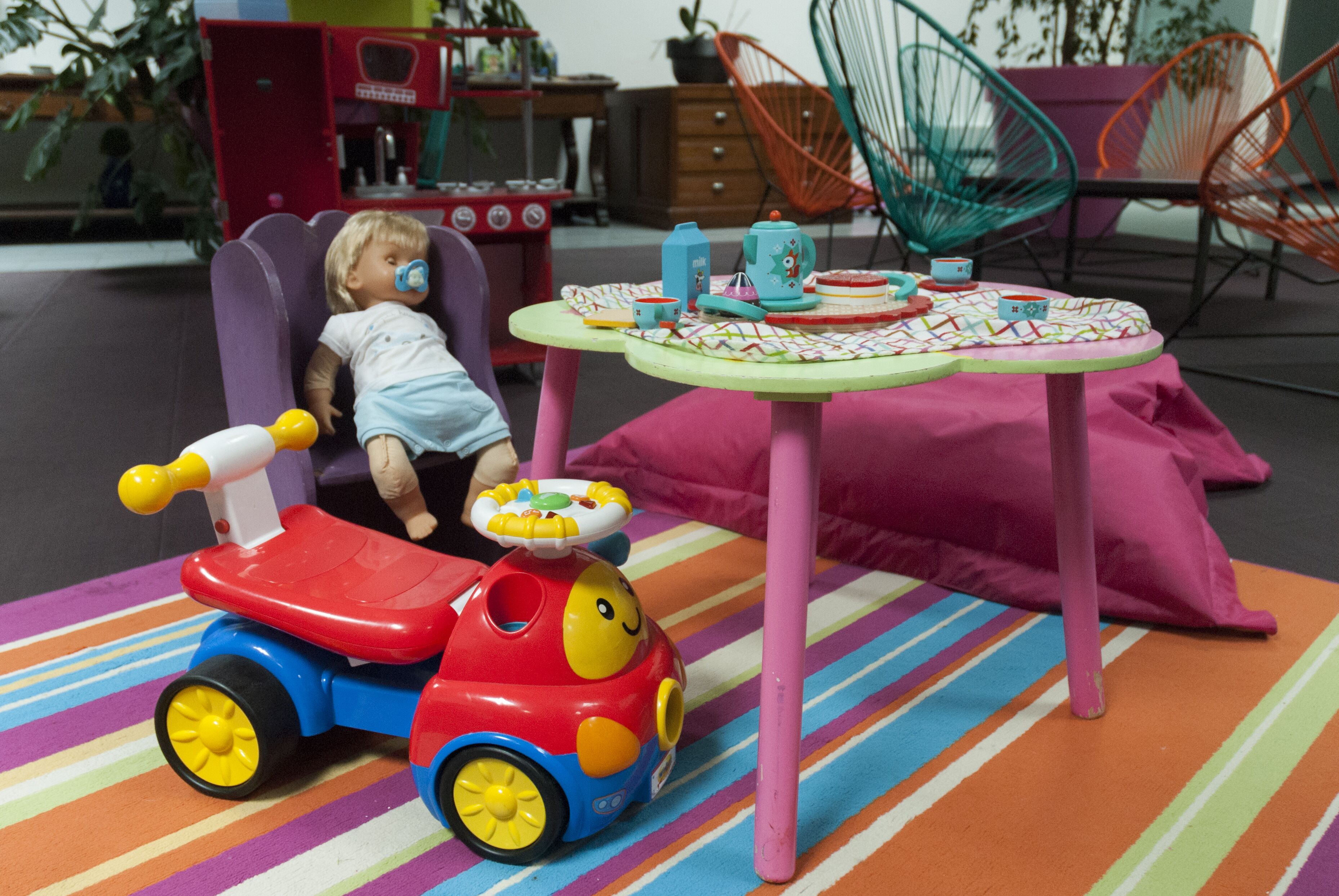 Una muñeca sentada en un coche de juguete, con una mesa y sillas al fondo.