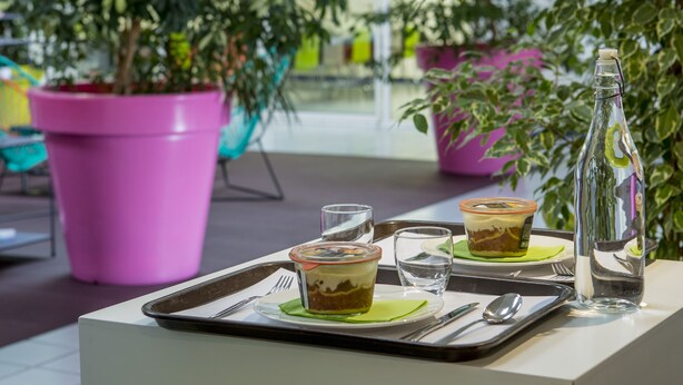 A table set with a dessert, a bottle, and a green chair in a modern indoor setting with large potted plants.