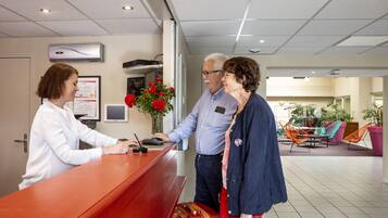 Un mostrador de recepción con una recepcionista, un hombre y una mujer de pie. Se observa un jarrón con flores rojas, un monitor y una puerta al fondo.