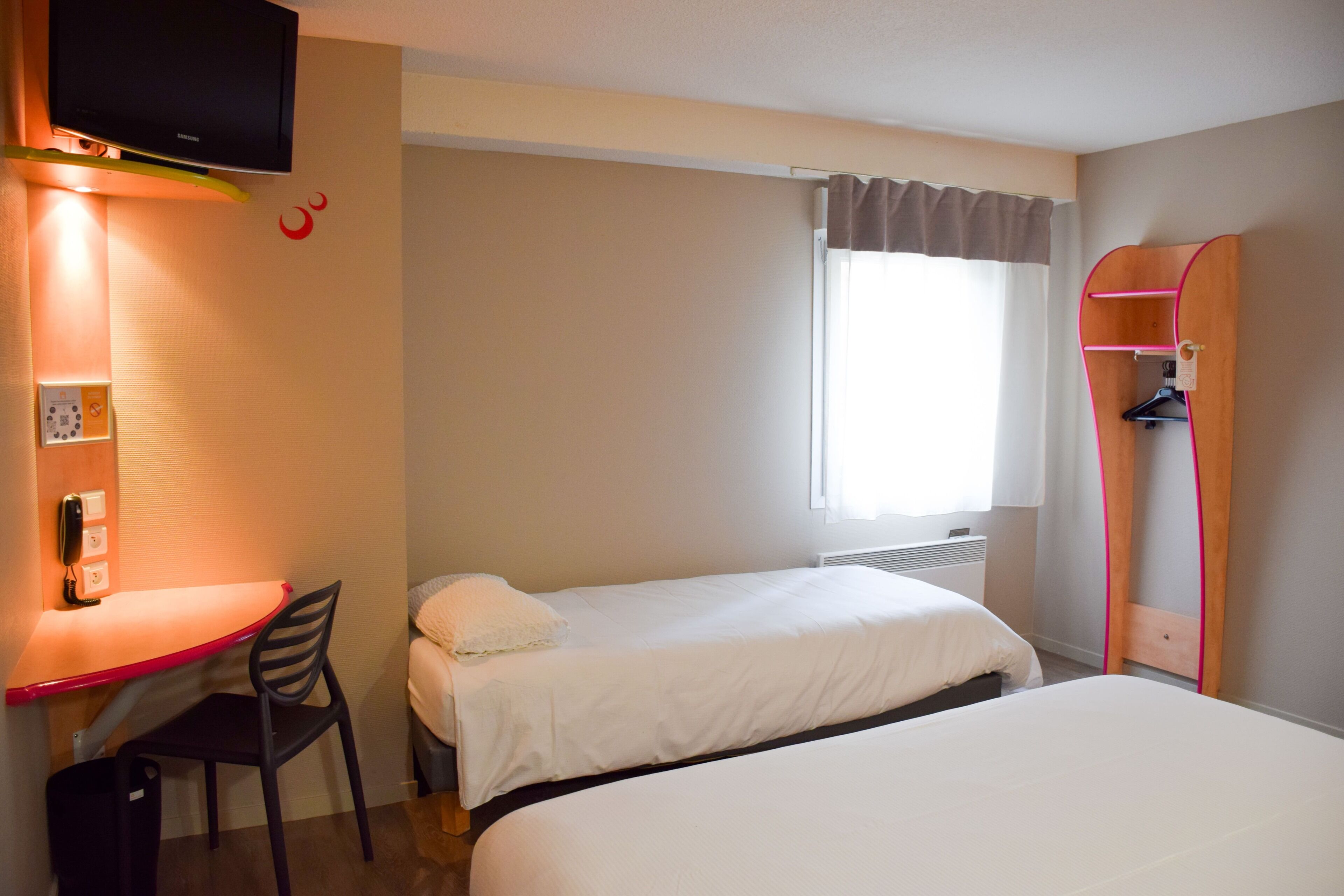 A hotel room with two single beds, a desk with a chair, a television mounted on the wall, and a window with curtains.
