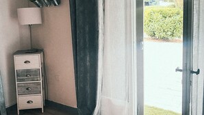 Una habitación con piso de madera, una mesita de noche blanca con cajones, una lámpara y una cortina.