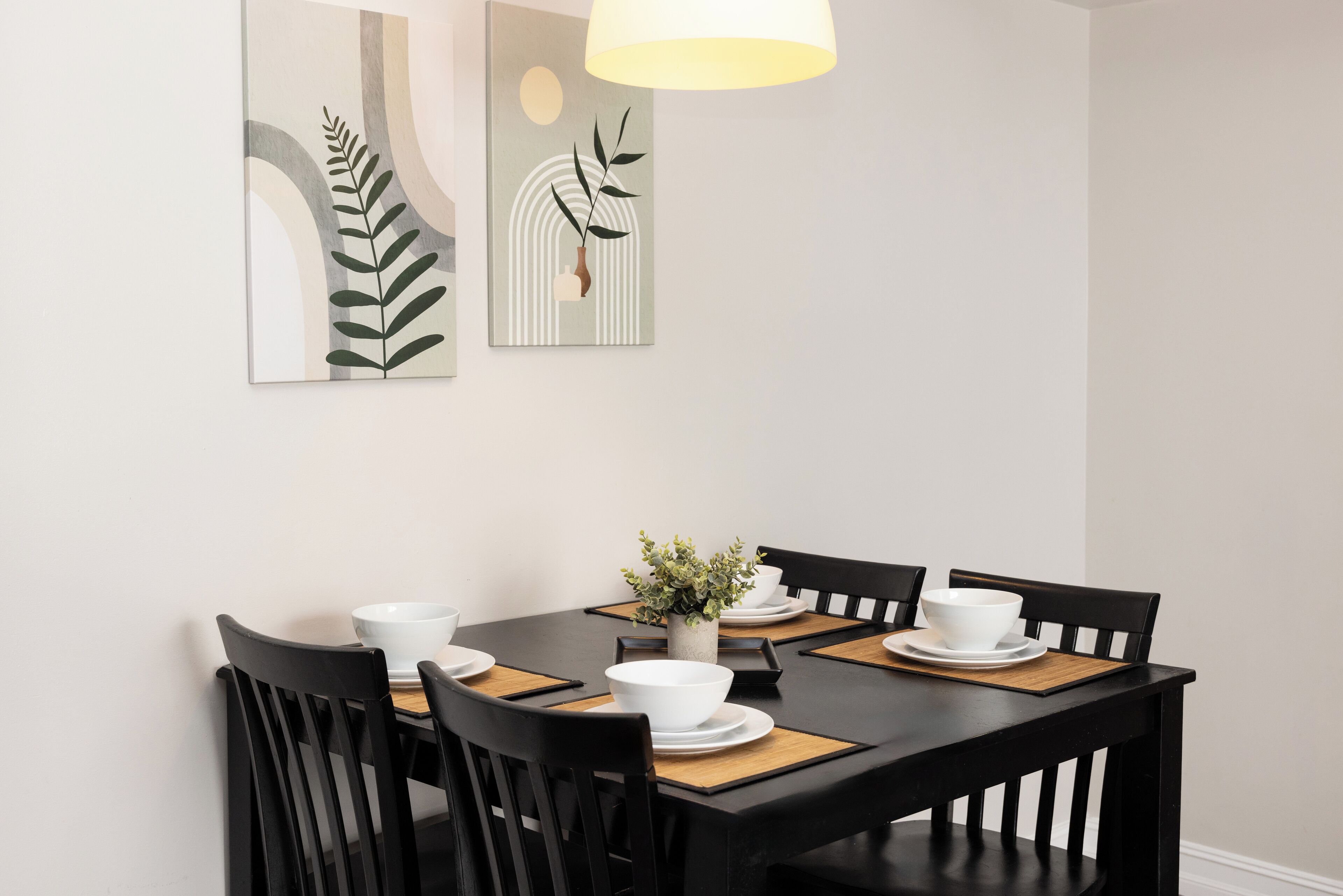 Un comedor con una mesa negra, cuencos blancos y sillas negras. Se observan dos obras de arte colgadas en la pared sobre la mesa.