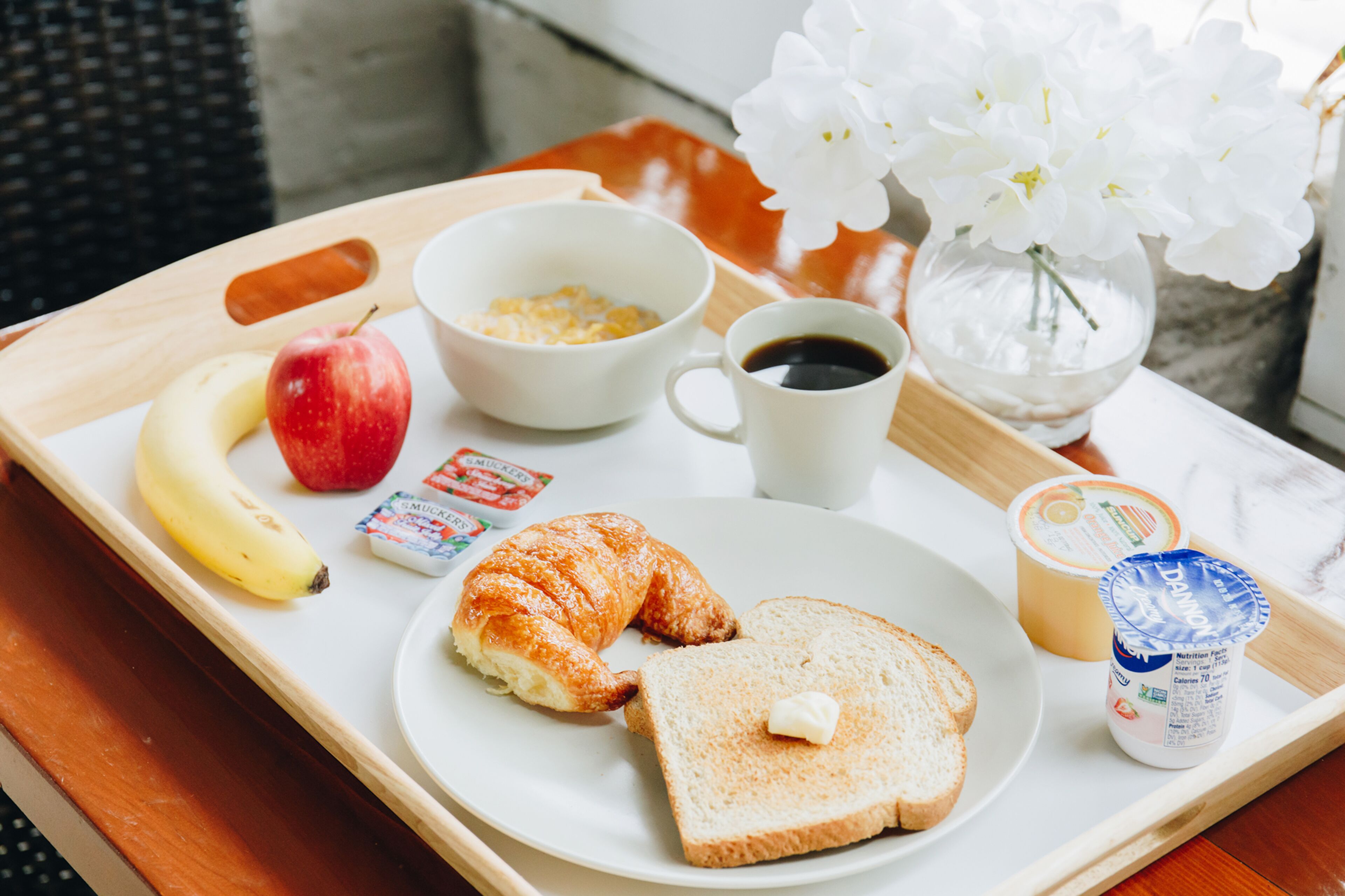 A tray with breakfast items including a croissant, two slices of toast with butter, a banana, an apple, a cup of coffee, a bowl of cereal, and a container of yogurt.