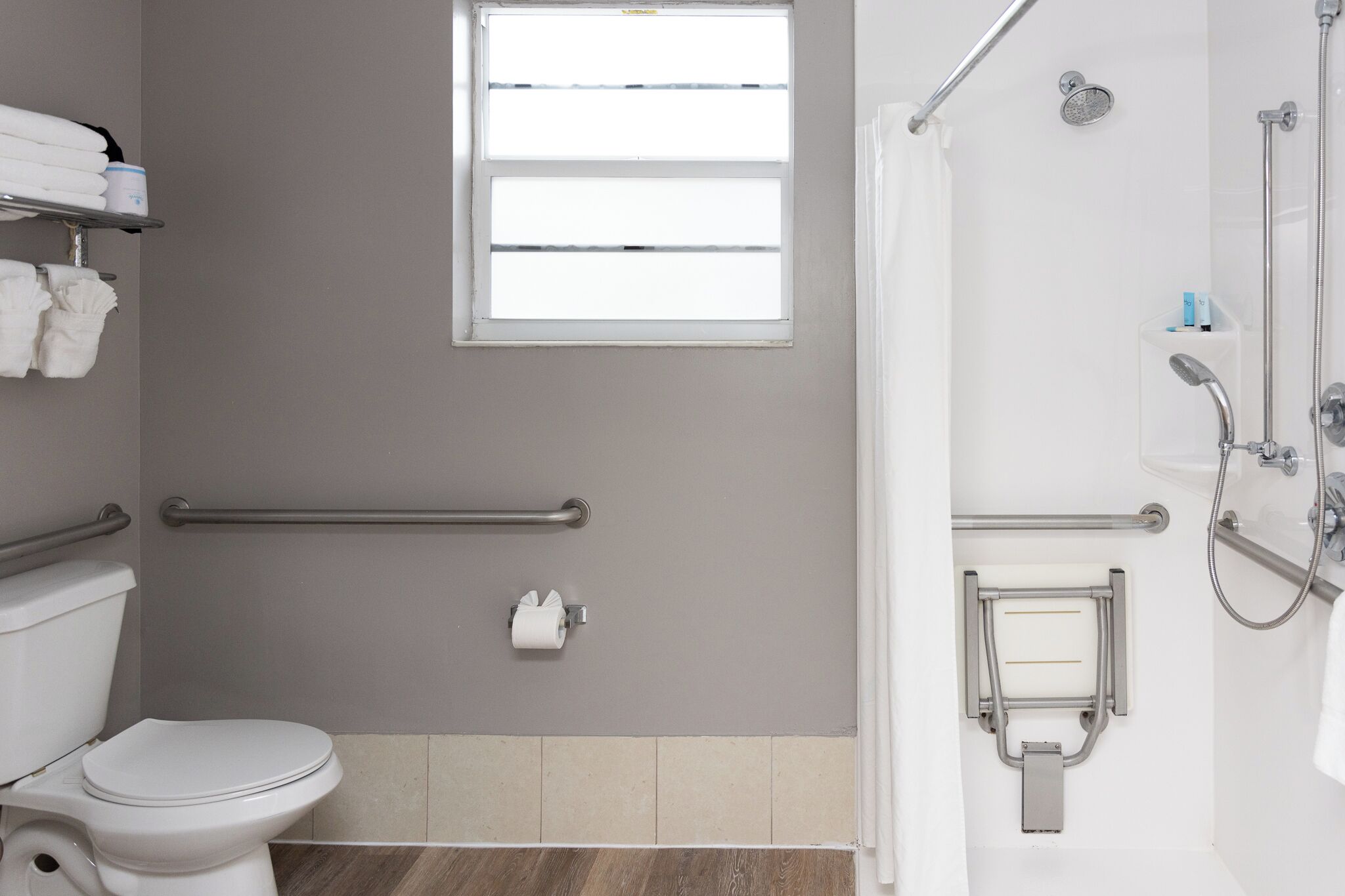 A modern bathroom with a toilet, grab bars, a window, and a shower area.