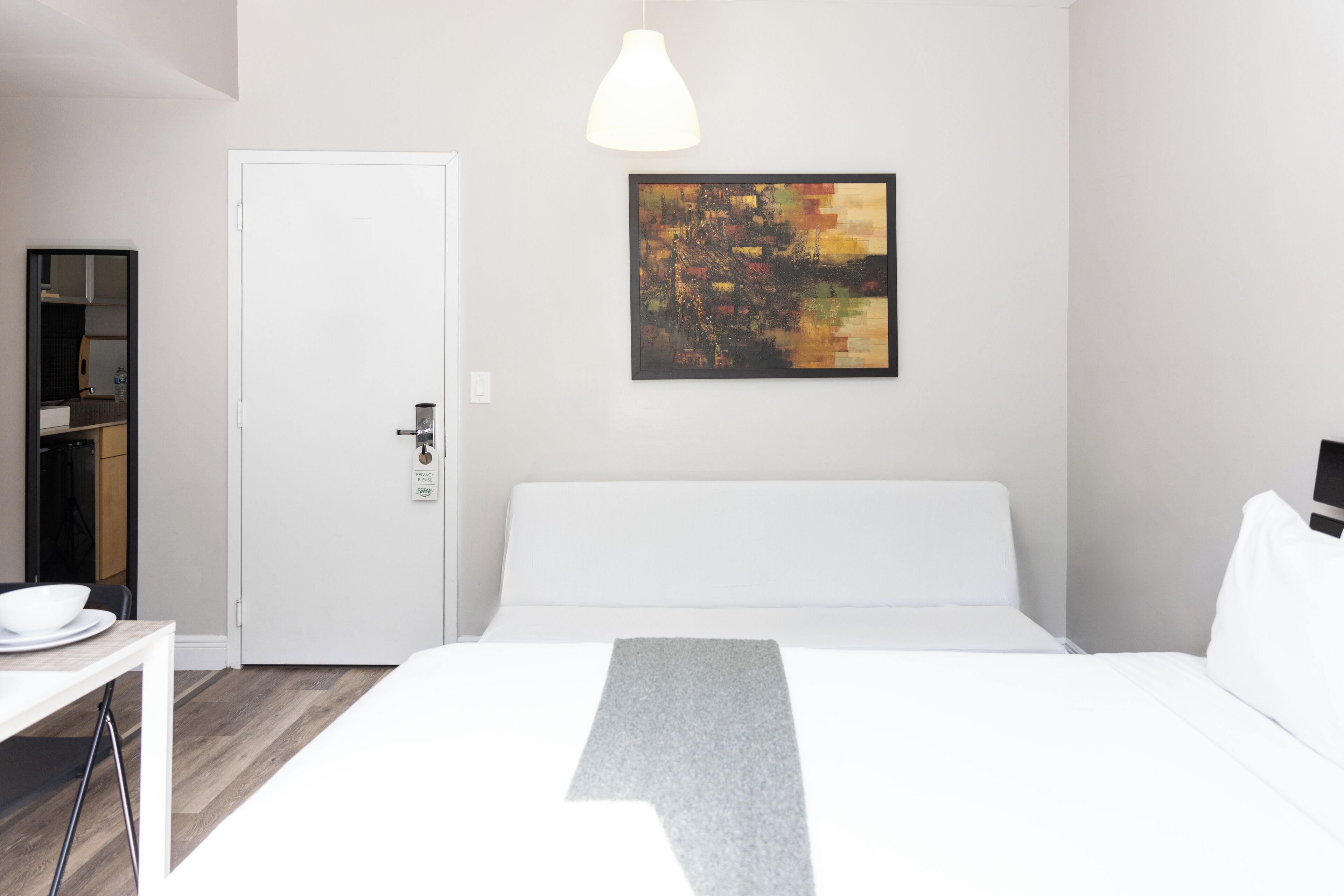 A hotel room with a bed, a desk, a chair, a white door, and a painting on the wall.