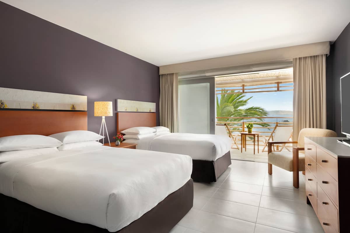 A hotel room with two beds, a desk, a chair, and a balcony with a view.