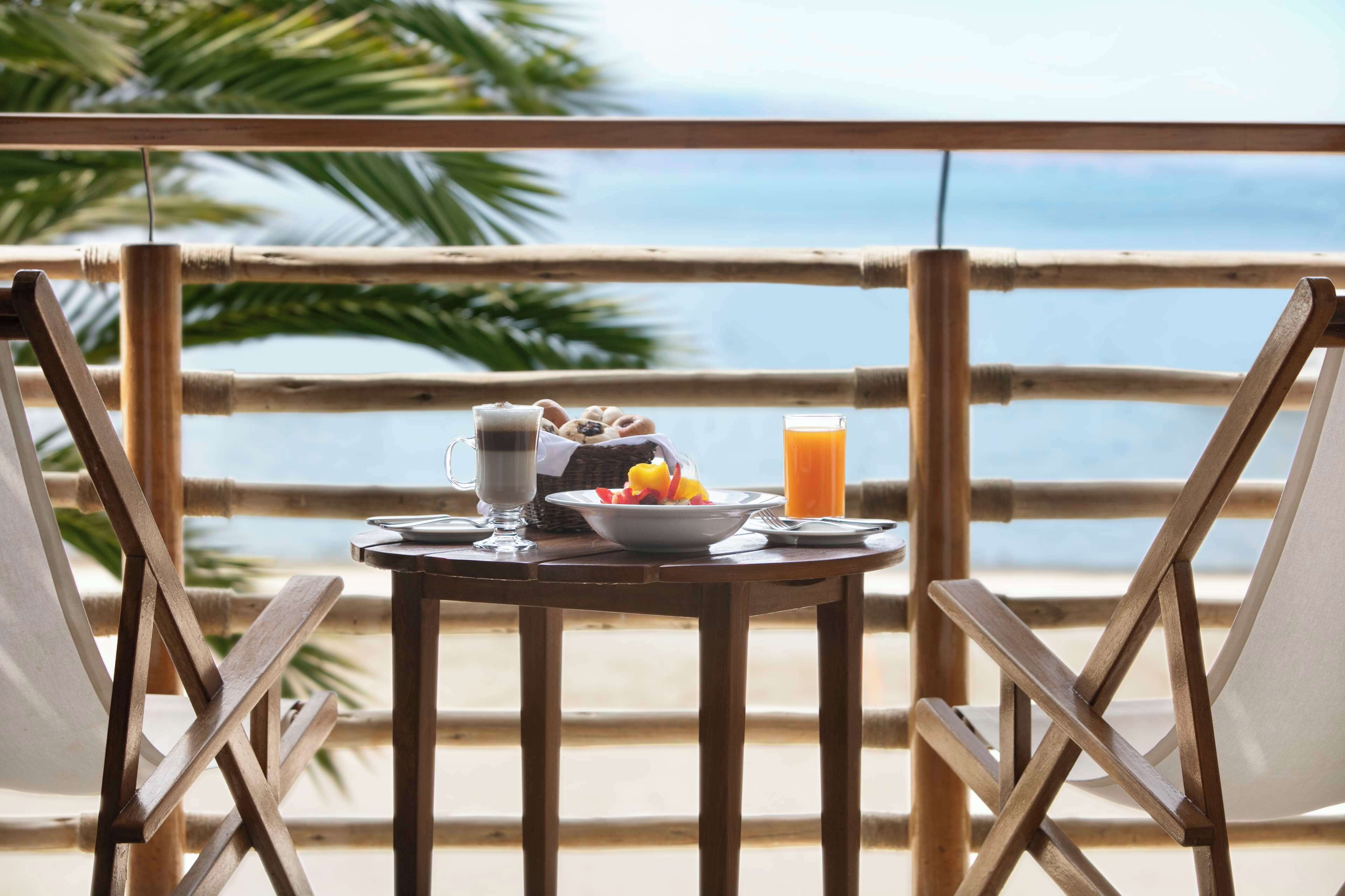 Una mesa con alimentos para el desayuno y un vaso de jugo de naranja en un balcón con vista a la playa.