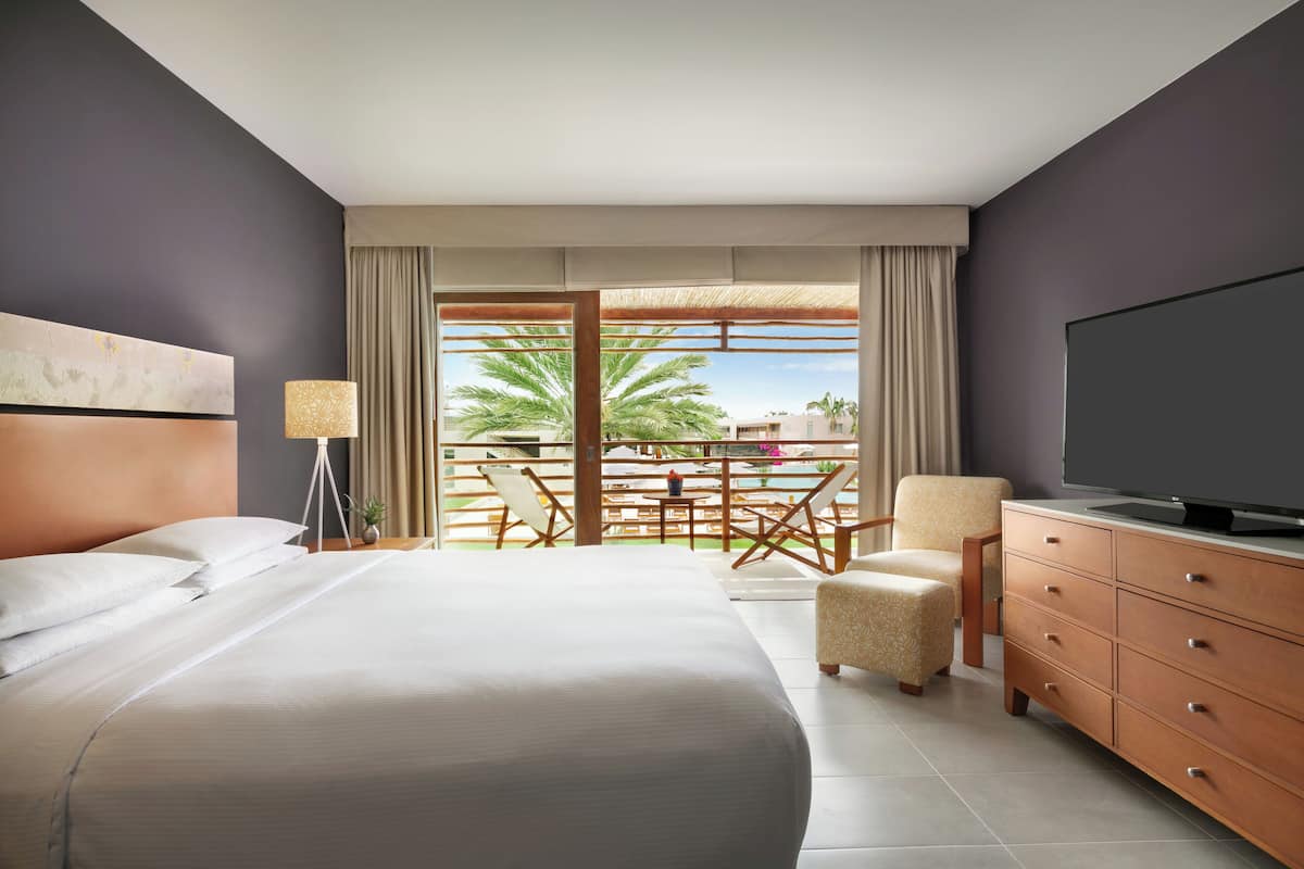A hotel room with a large bed, a TV, a dresser, and a balcony with outdoor furniture.