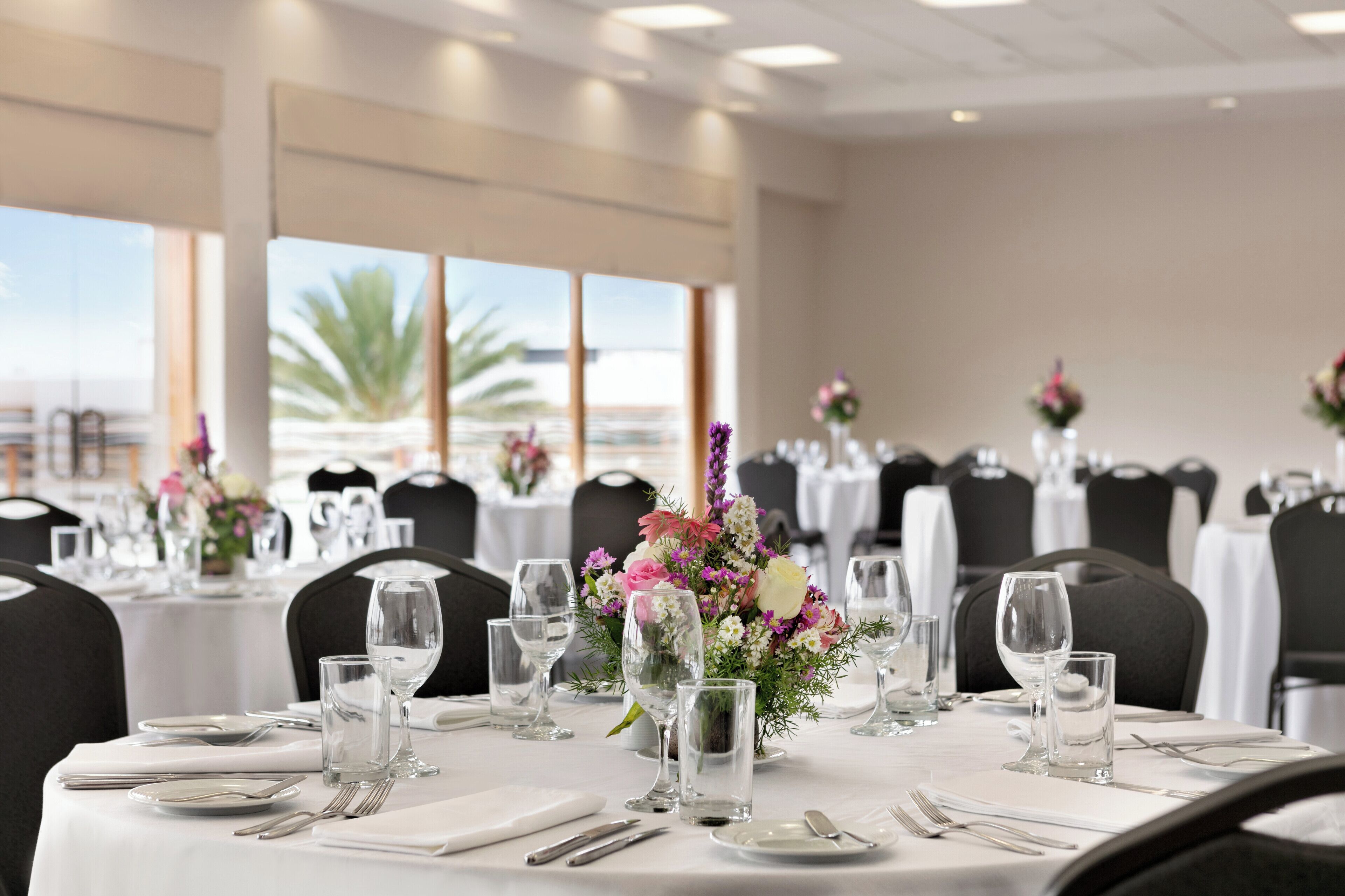 A dining area with tables set for a meal, featuring floral centerpieces and a view of the ocean through large windows.