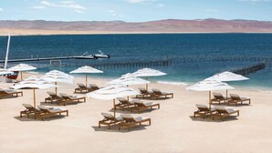 Ein Strand mit Liegestühlen und Sonnenschirmen, ein Steg, der sich ins Wasser erstreckt, und Berge im Hintergrund.