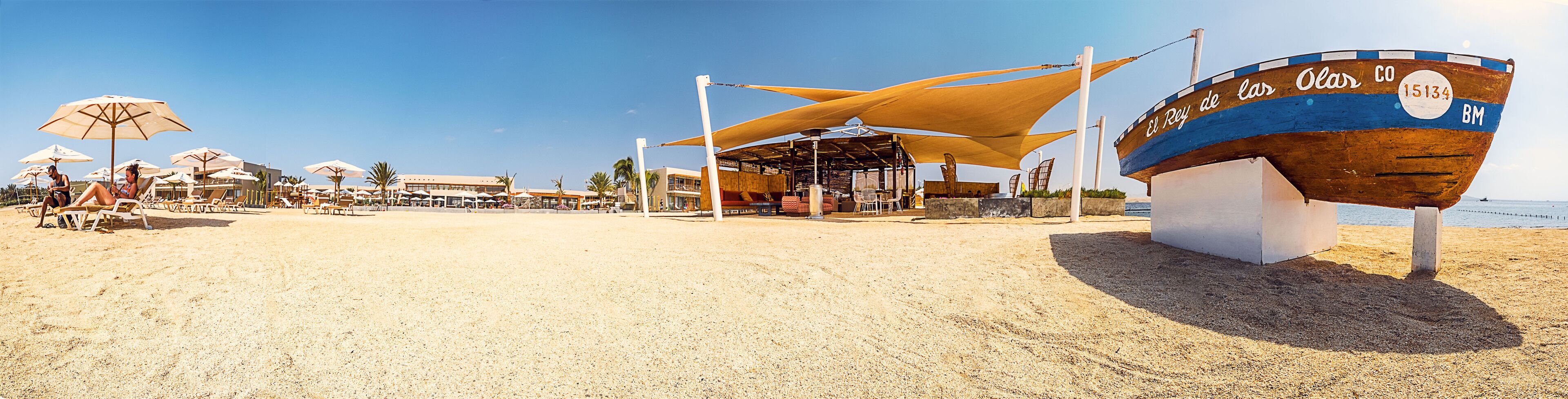 A beach with sun loungers and umbrellas, a boat named 'El Rey de las Olas', and a covered area with tables and chairs.
