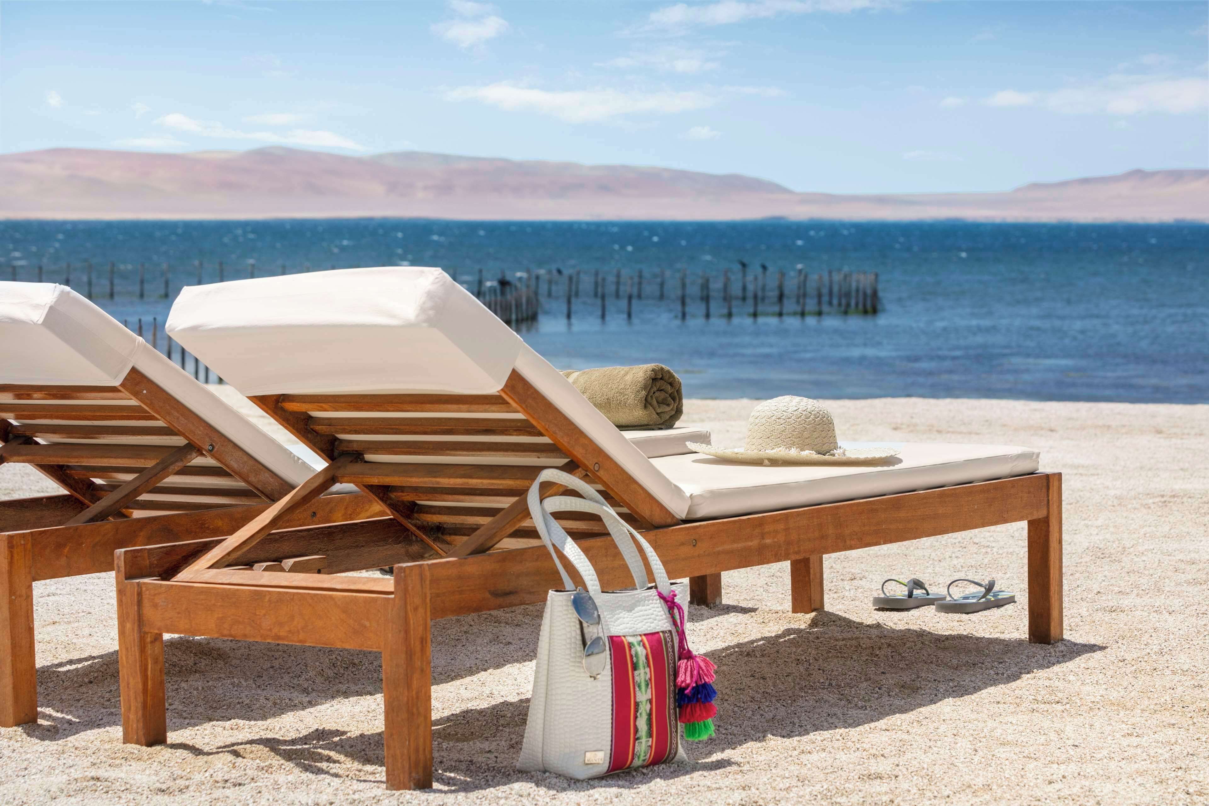 Dos sillones de madera con cojines blancos sobre una playa arenosa, una toalla enrollada, un bolso colorido y un par de chanclas.