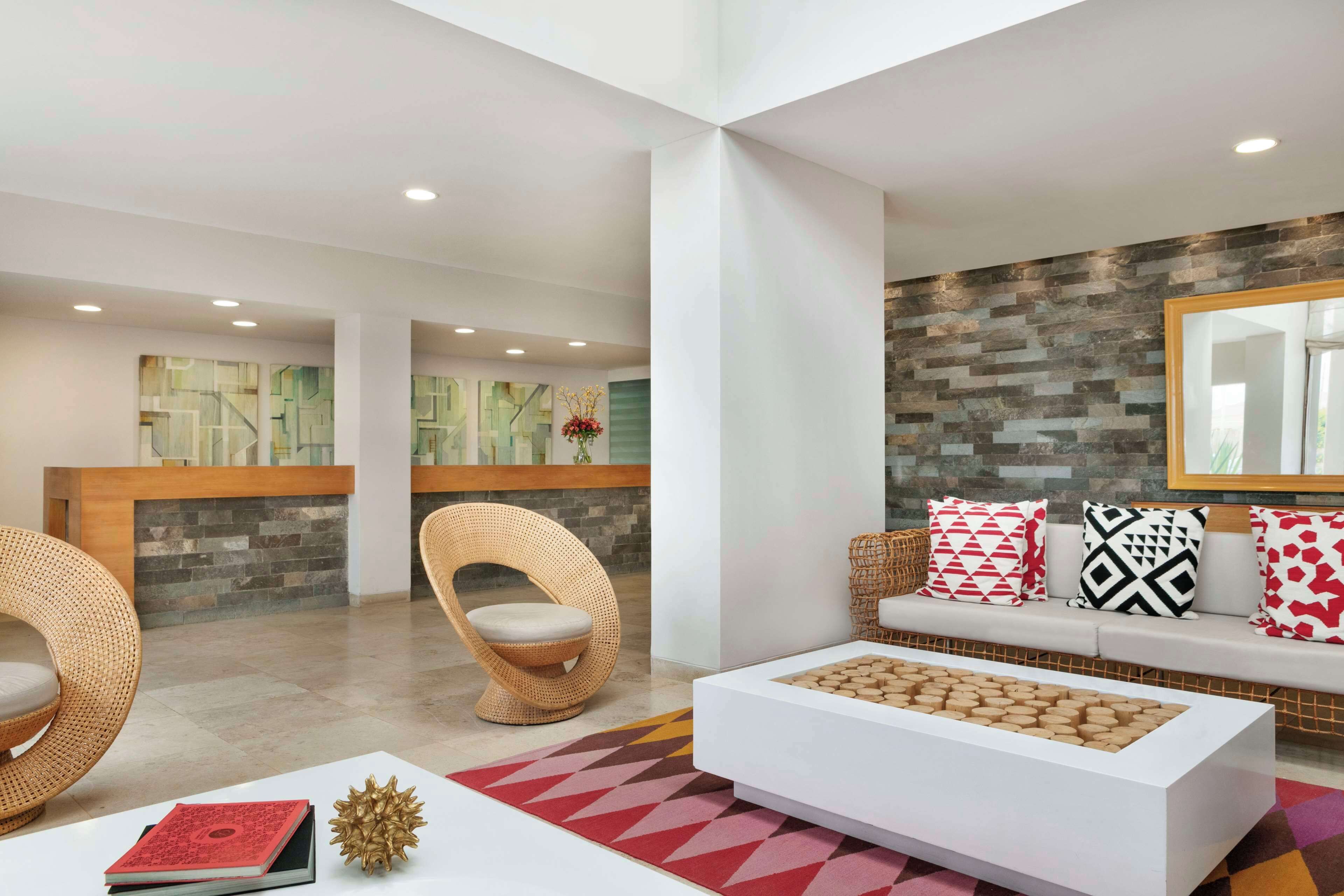 A modern lobby with a reception desk, wicker chairs, a white sofa with patterned cushions, and a coffee table with wooden blocks.