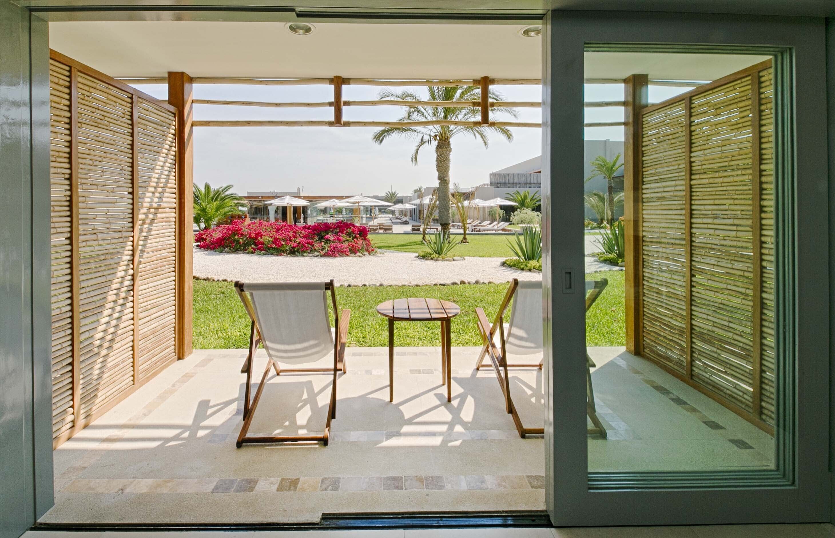 Un patio con mesa y sillas, vista a un jardín con palmeras y flores, y un edificio al fondo.