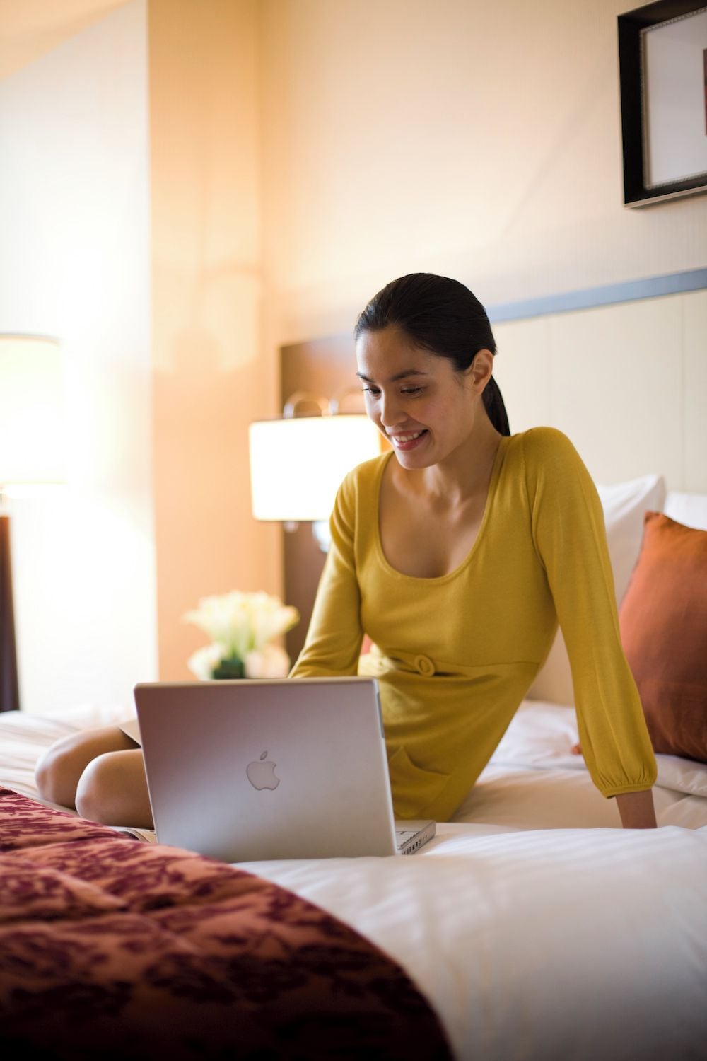 A person sitting on a bed with a laptop, in a room with a lamp and a framed picture on the wall.