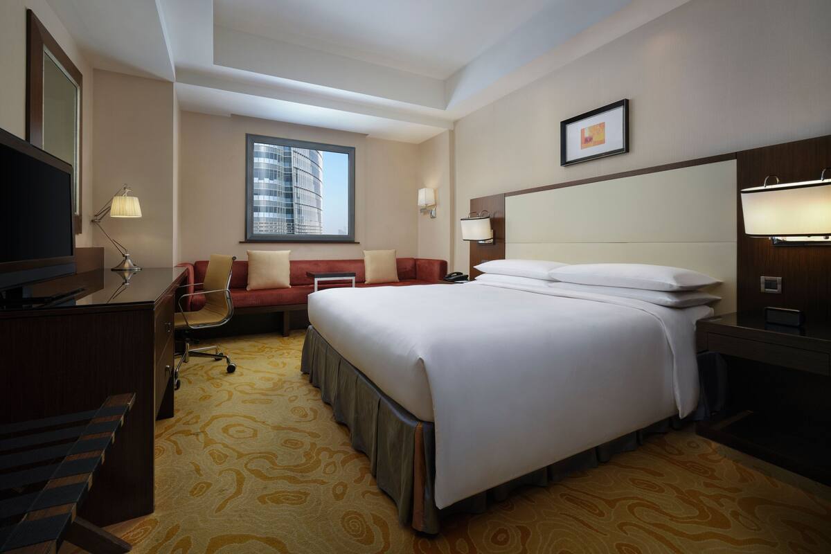 Ein Hotelzimmer mit einem großen Bett, einem Schreibtisch mit Computer, einem Sessel, einer Couch und einem Fenster mit Blick auf ein Gebäude.