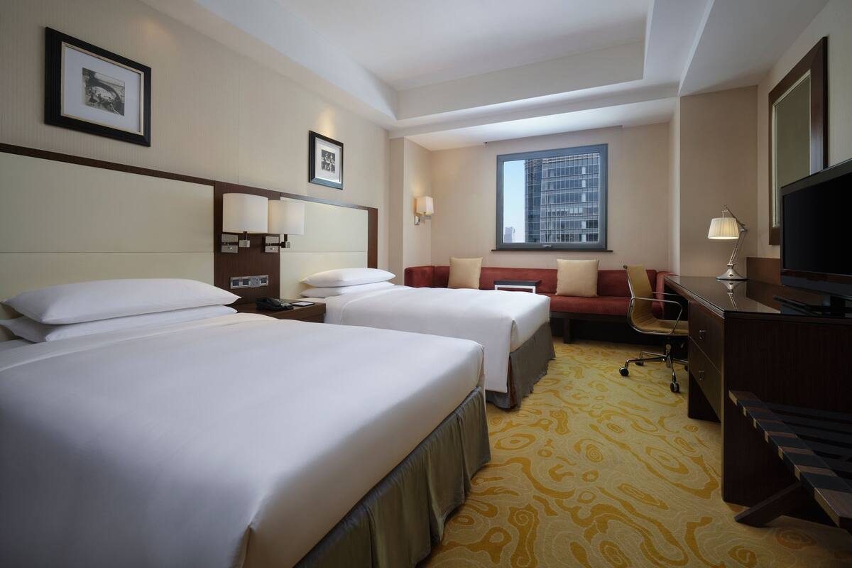 Ein Hotelzimmer mit zwei Betten, einem Schreibtisch, einem Stuhl, einem Fernseher und einem Fenster mit Blick auf die Stadt.