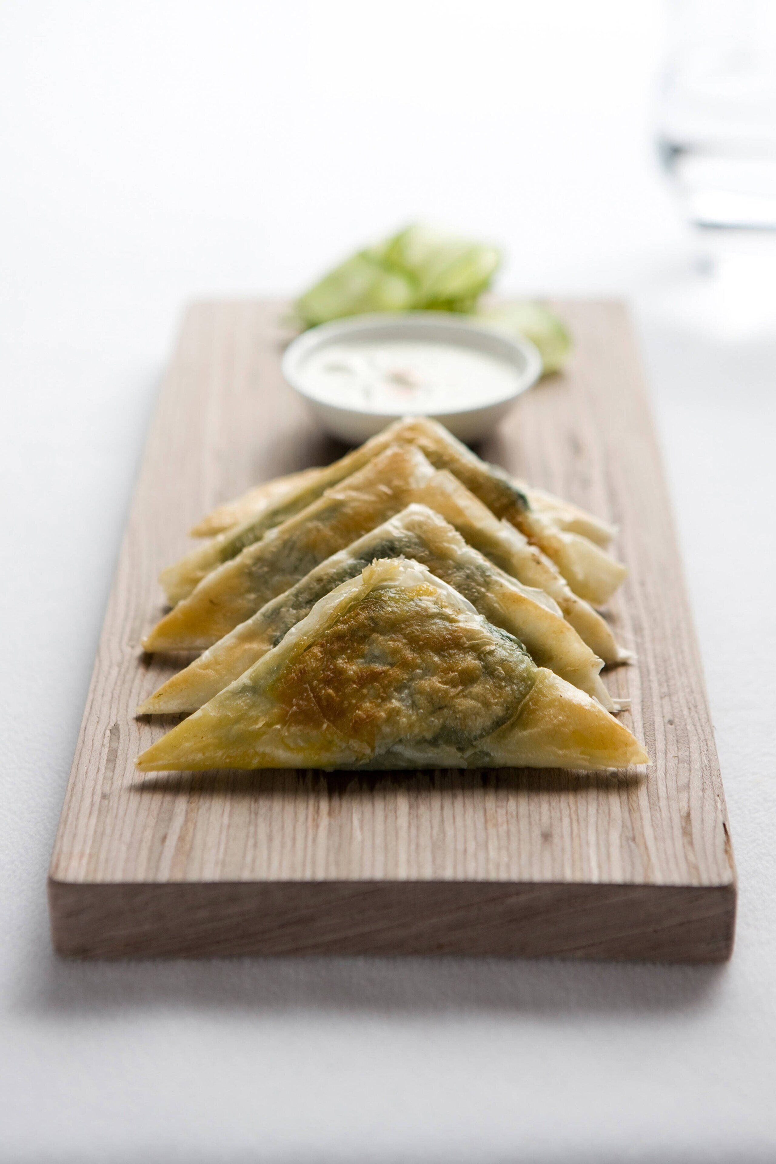 A wooden serving board with triangular pastries and a small bowl of dipping sauce.