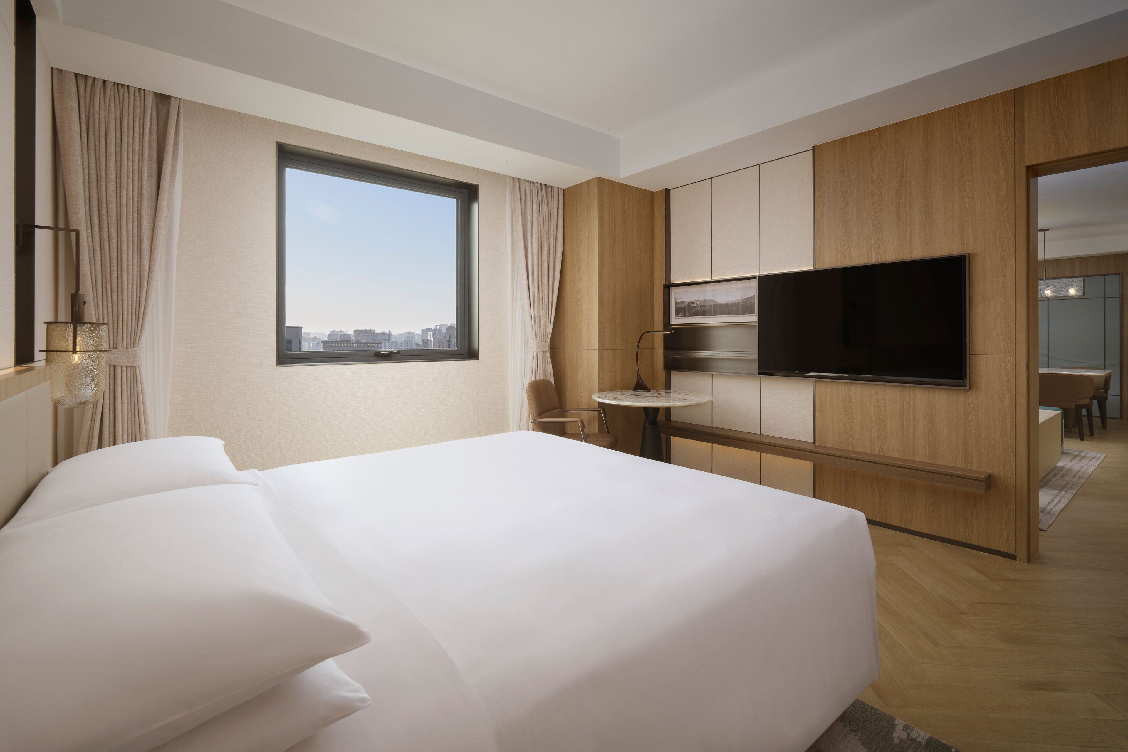 A modern hotel room with a large bed, a TV mounted on the wall, and a city view through the window.