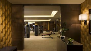 A hotel lobby with a reception desk, a bench, a wall-mounted lamp, and exercise equipment in the background.