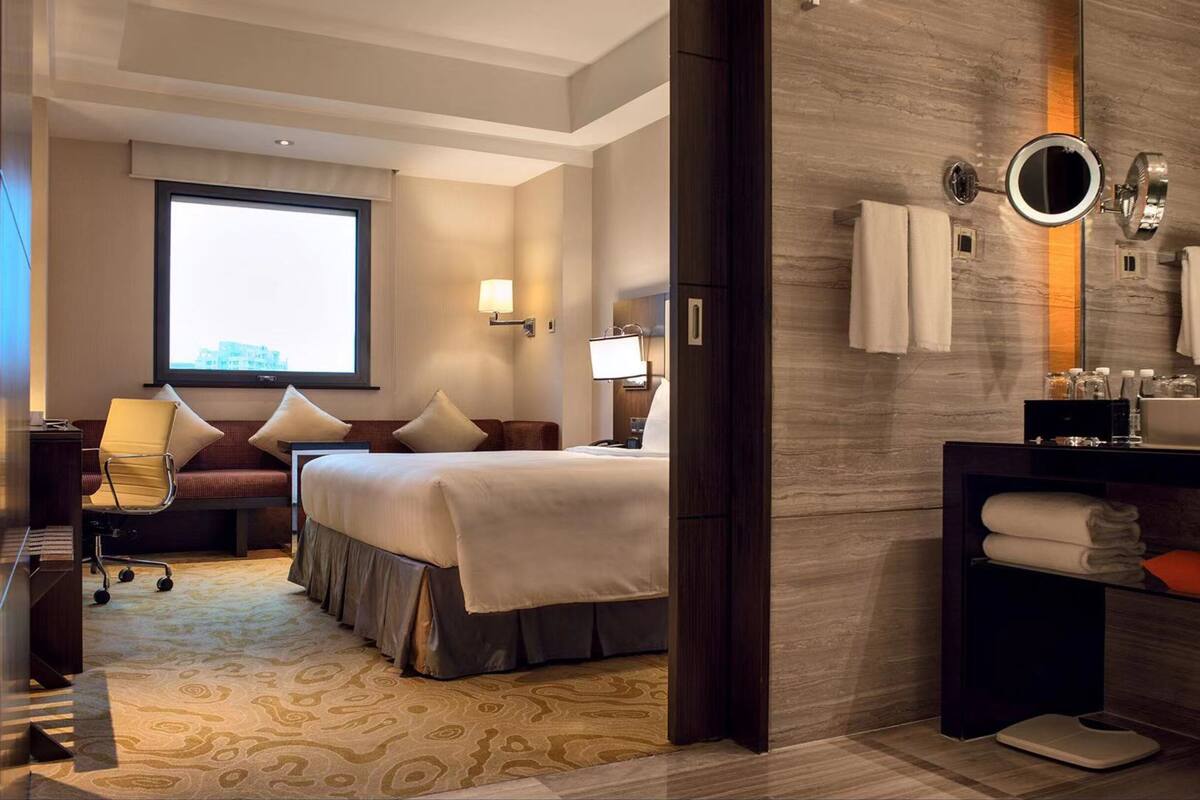 Ein Hotelzimmer mit Bett, Nachttisch, Lampe und einem großen Fenster.