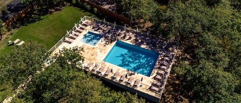 An aerial view of a rectangular swimming pool with a tiled deck, surrounded by lounge chairs and umbrellas, set in a wooded area.