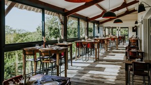Un área de comedor al aire libre con mesas y sillas de madera, ventanas amplias y lámparas colgantes.