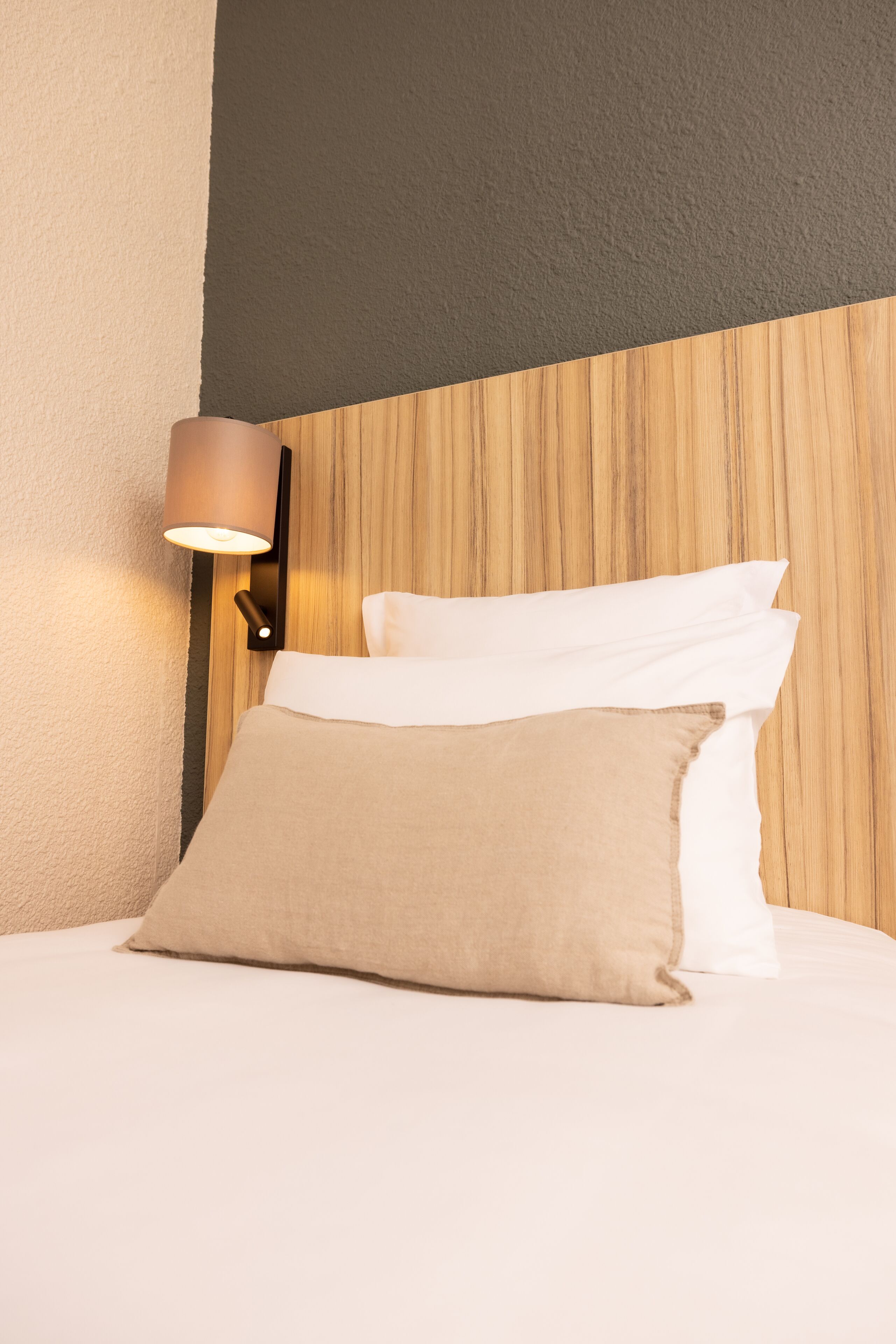 Una cama bien hecha con almohadas blancas y beige, un cabecero de madera y una lámpara fijada a la pared.