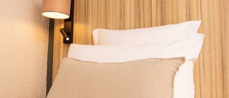 A neatly made bed with white and beige pillows, a wooden headboard, and a wall-mounted lamp.
