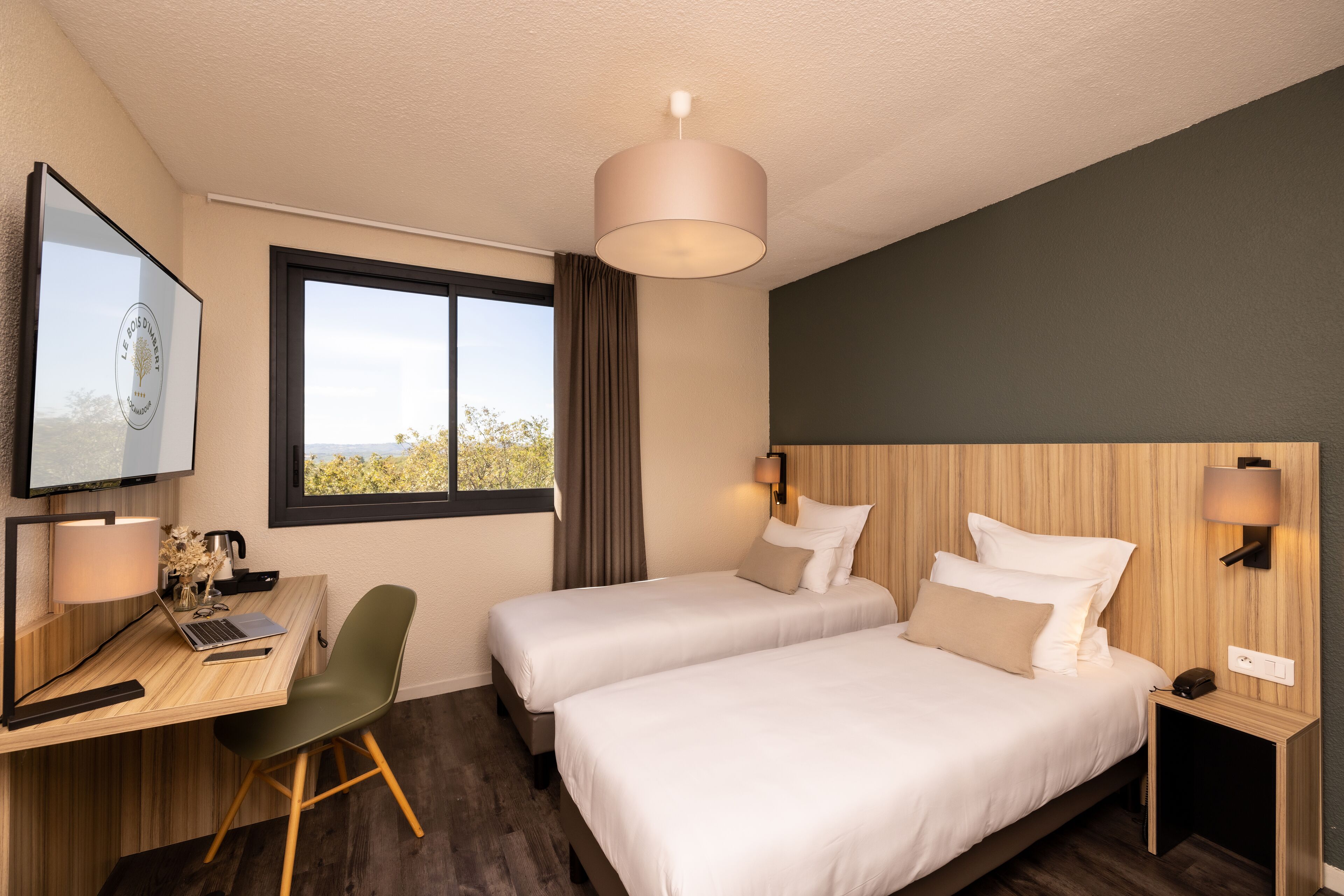 A hotel room with two beds, a desk with a laptop, a chair, a TV, and a window with a view.