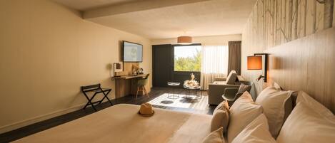A hotel room with a large bed, a sofa, a desk, and a chair. There is a window with curtains and a view of trees.