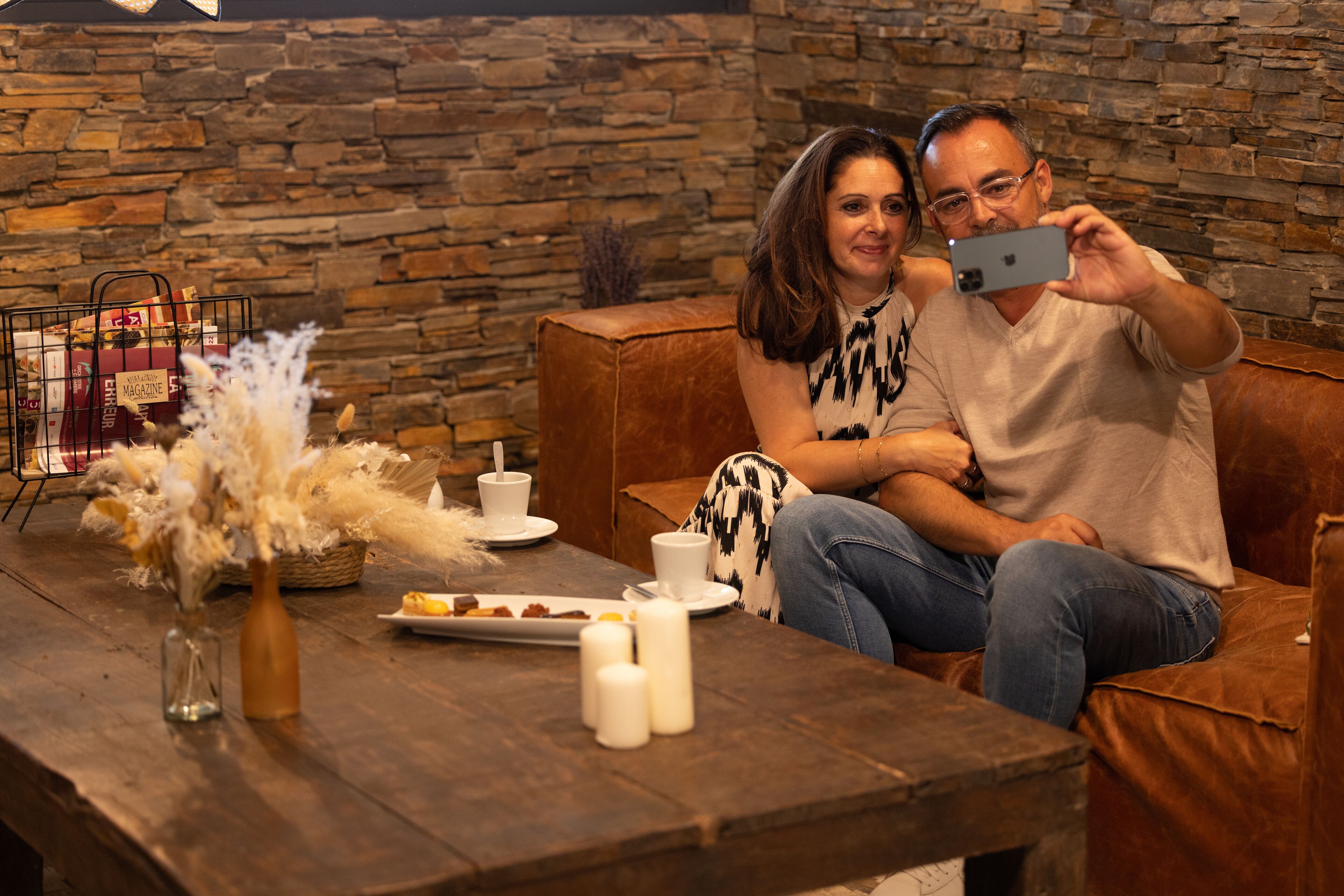A couple sitting on a leather couch in a room with a stone wall, a wooden table with a vase of dried flowers, and a magazine rack.