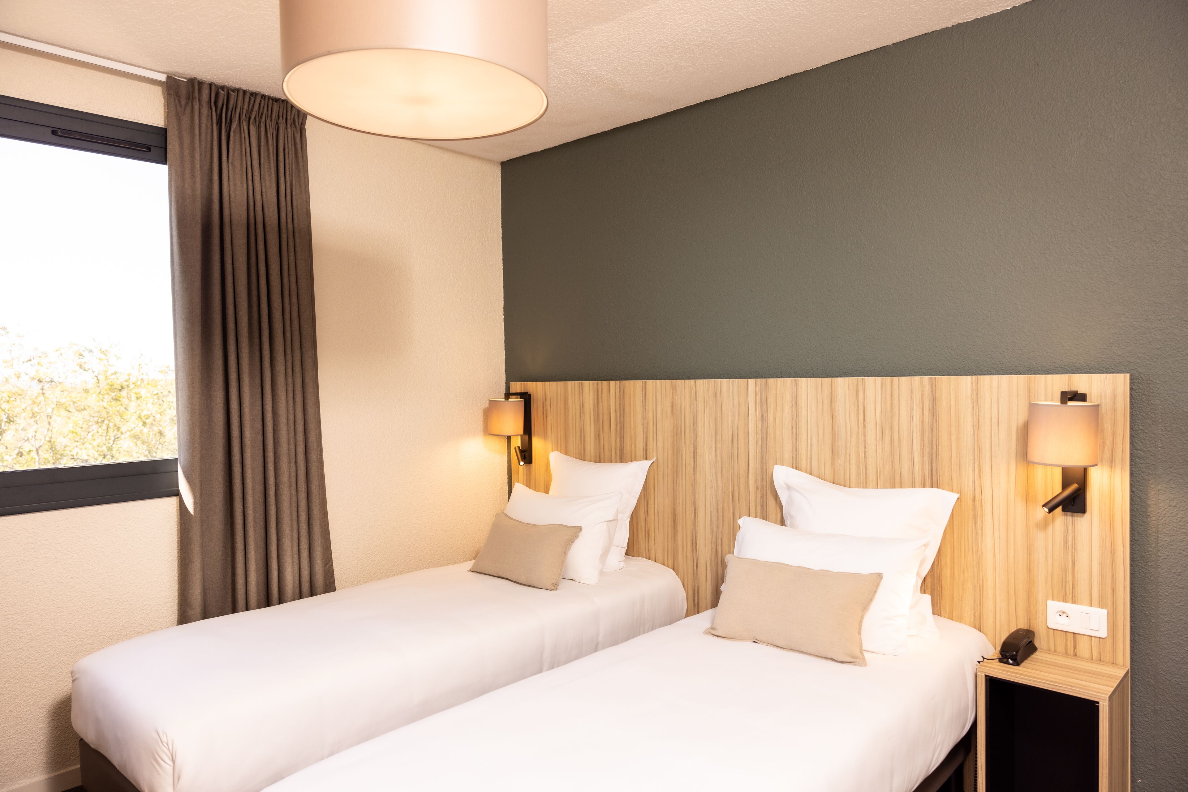 Una habitación de hotel con dos camas, un ventanal con cortinas y lámparas fijadas a la pared.